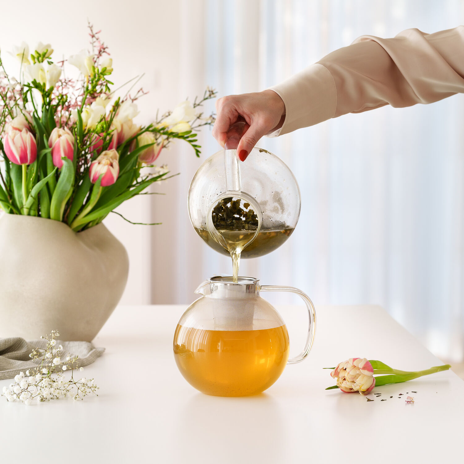 Frau gießt frisch aufgebrühten Tee aus einer Glaskanne mit integriertem Sieb in eine bauchige Teekanne neben Frühlingsblumen auf einem Tisch