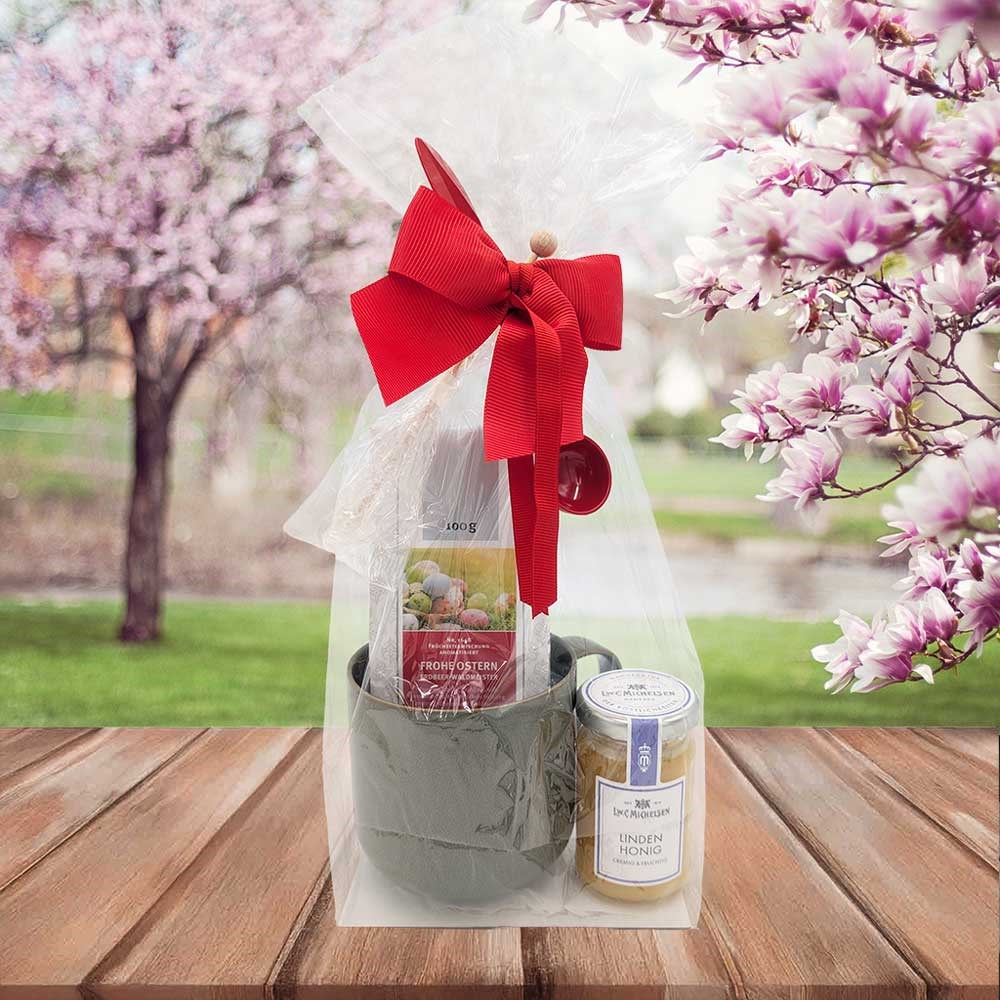 Geschenkset mit Keramiktasse, Ostertee und Glas Lindenhonig, in Folie mit roter Schleife verpackt, auf Holztisch vor blühenden Frühlingsbäumen