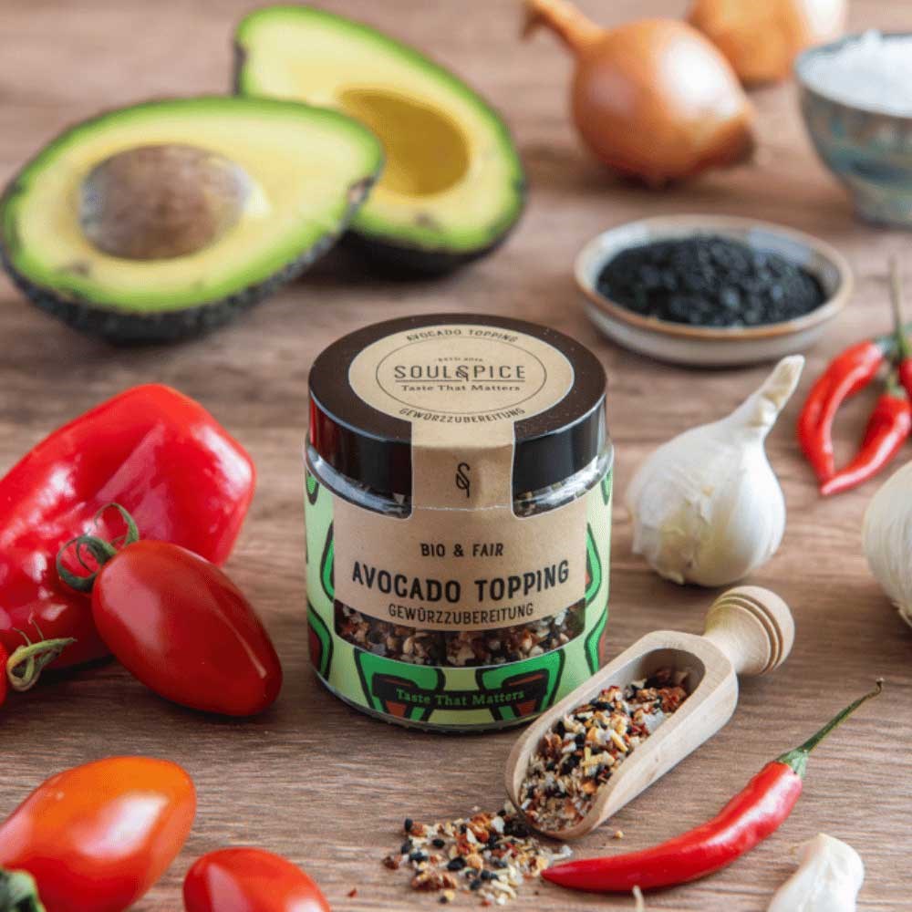 Jar of SoulSpice organic & fair avocado topping spice blend surrounded by avocado, tomatoes, peppers, garlic and chilli on wooden table