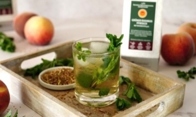 Glas mit Pfirsich-Minz-Cocktail auf Tablett, daneben Kräuter, Gewürze, frische Pfirsiche und Produktverpackung