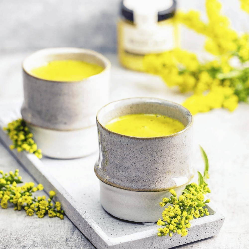 Two cups of golden milk turmeric latte in modern ceramic mugs on a tray, decorated with yellow blossoms