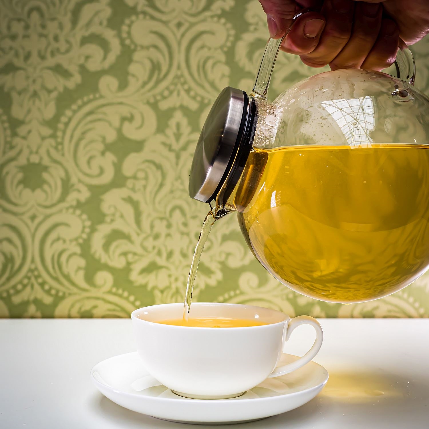 Hand gießt heißen Kräutertee aus Glaskanne in Tasse, Teekanne, Kaffeetee, Glas-Kanne, Teewasser, Kaffeetasse