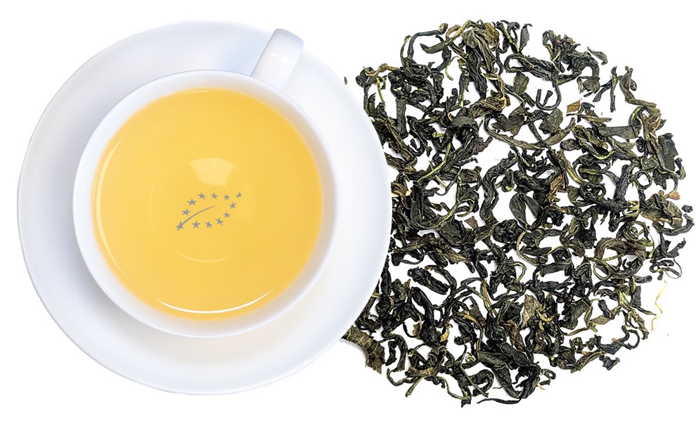 White cup with light yellow green tea next to loose green tea leaves on white background