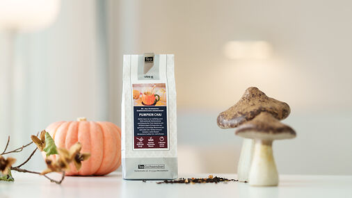 Teetüte mit Pumpkin Chai Tee auf einem hellen Untergrund, daneben stehen Dekopilze und ein Dekokürbis