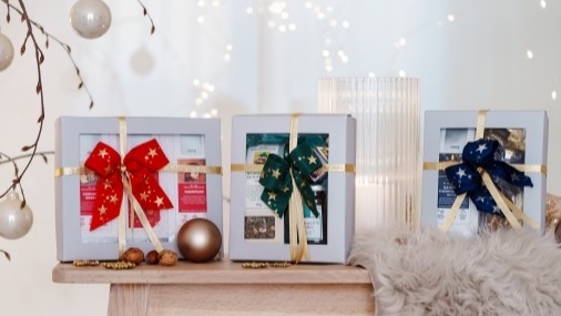 Drei weihnachtlich gestaltete Tee-Präsente in hellgrauen Kartonschachteln auf einem hellen Holztisch
