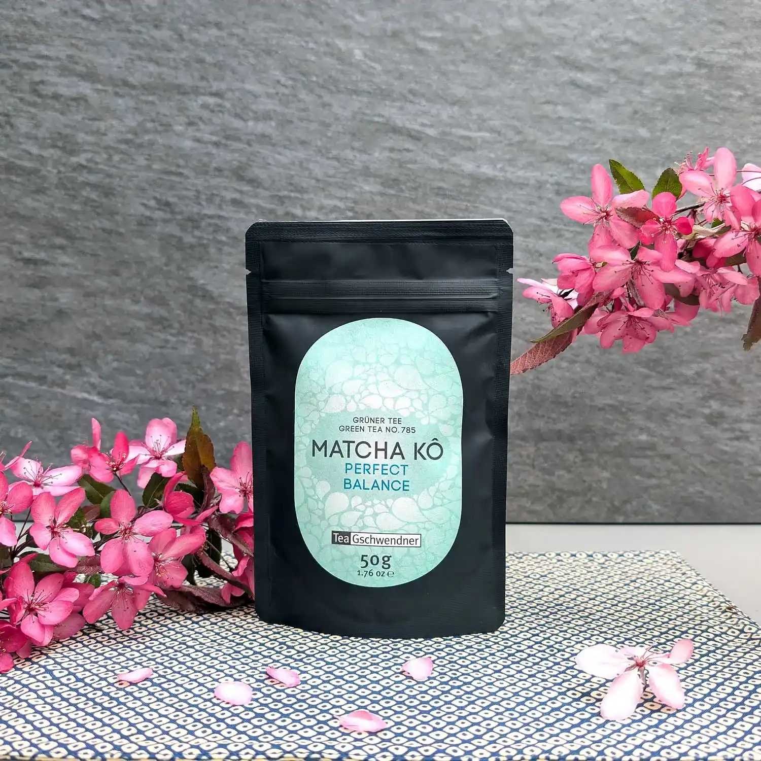 Black pouch of TeaGschwendner Matcha Kō Perfect Balance, green tea 50 g, styled with pink blossoms against a grey background
