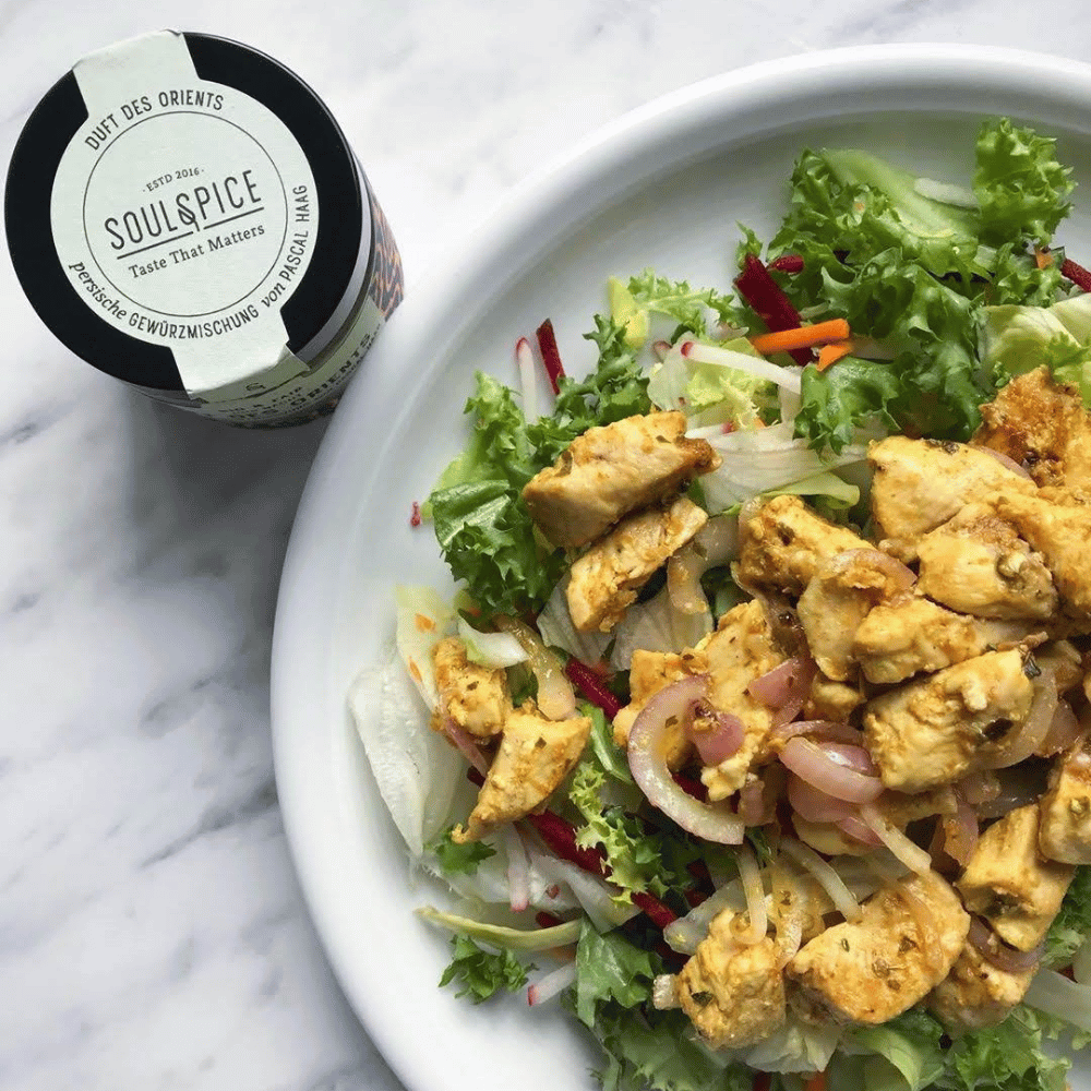 Assiette de salade verte mélangée, légumes et morceaux de poulet mariné à côté d’une boîte d’épices persanes SoulSpice