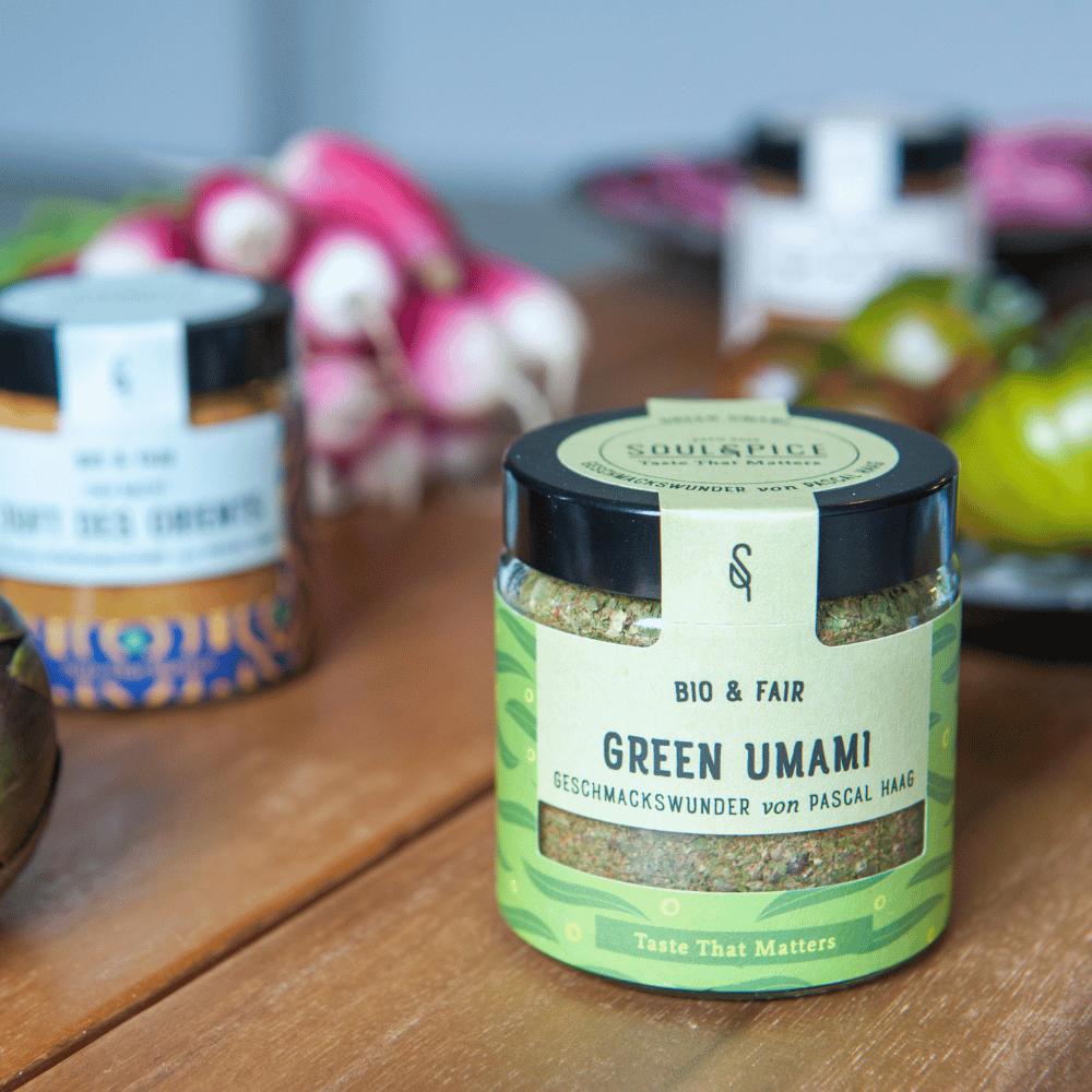 Glass jar of SOULSPICE Green Umami organic & fair spice blend by Pascal Haag on a wooden table, with other spices and fresh vegetables in the background