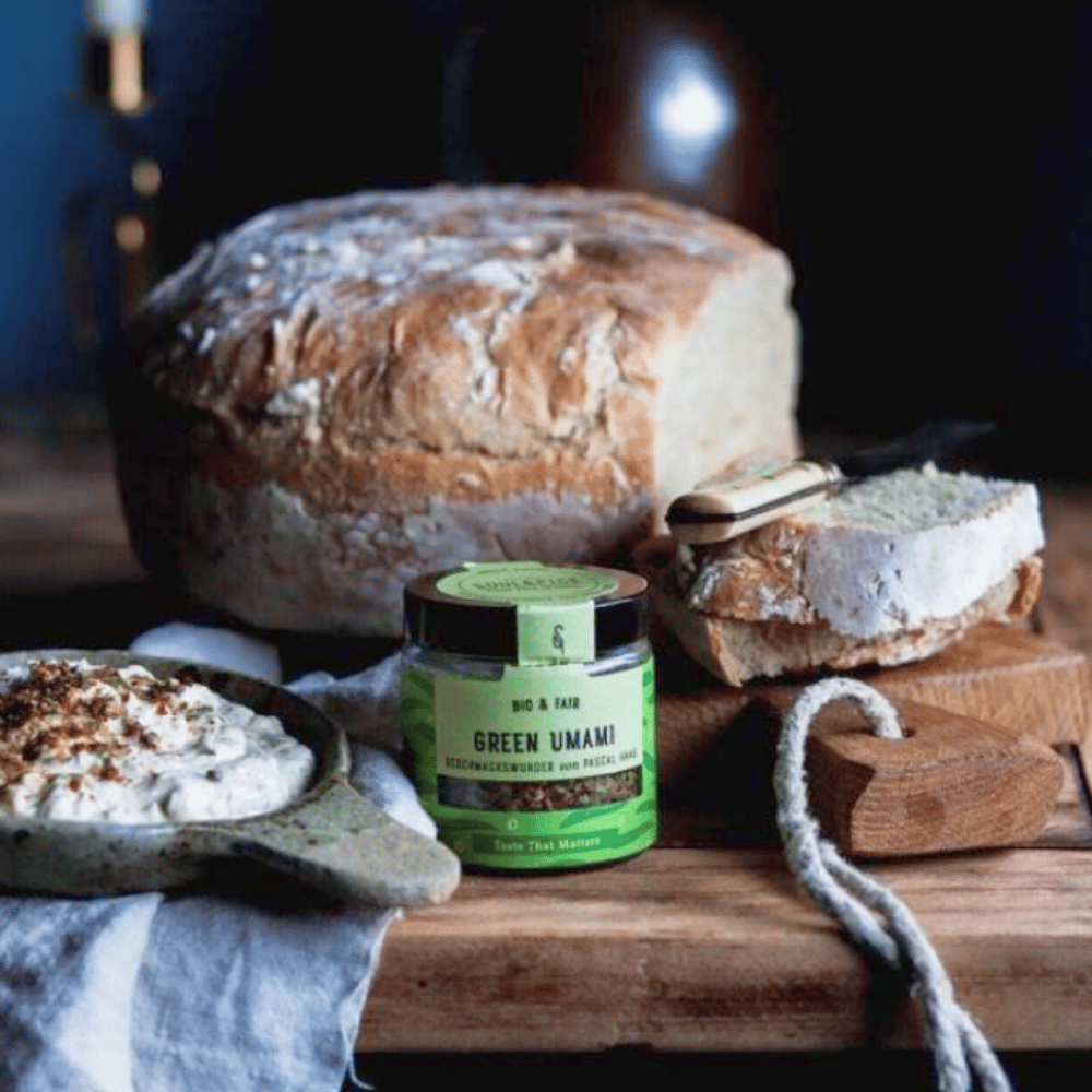 Jar of Green Umami organic seasoning blend in front of fresh farmhouse bread with spread on a rustic wooden table