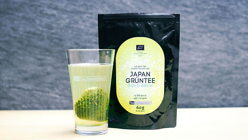 Grüner Tee mit Pyramidenbeutel in einem Glas, daneben eine schwarze Ziplocktüte mit der Aufschrift Japan Grüntee Cold Brew Bio