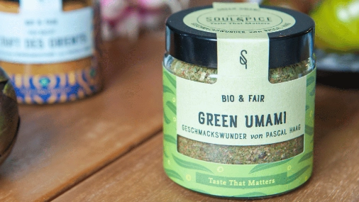 rundes Glas mit schwarzem Deckel, grüner Gewürzfülung und grünem Etikett mit der Aufschrift Green Umami auf einem Holztisch