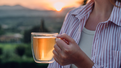 Teeglas mit gelbem Tee wird in einer Frauenhand gehalten vor einem Sonnenuntergang