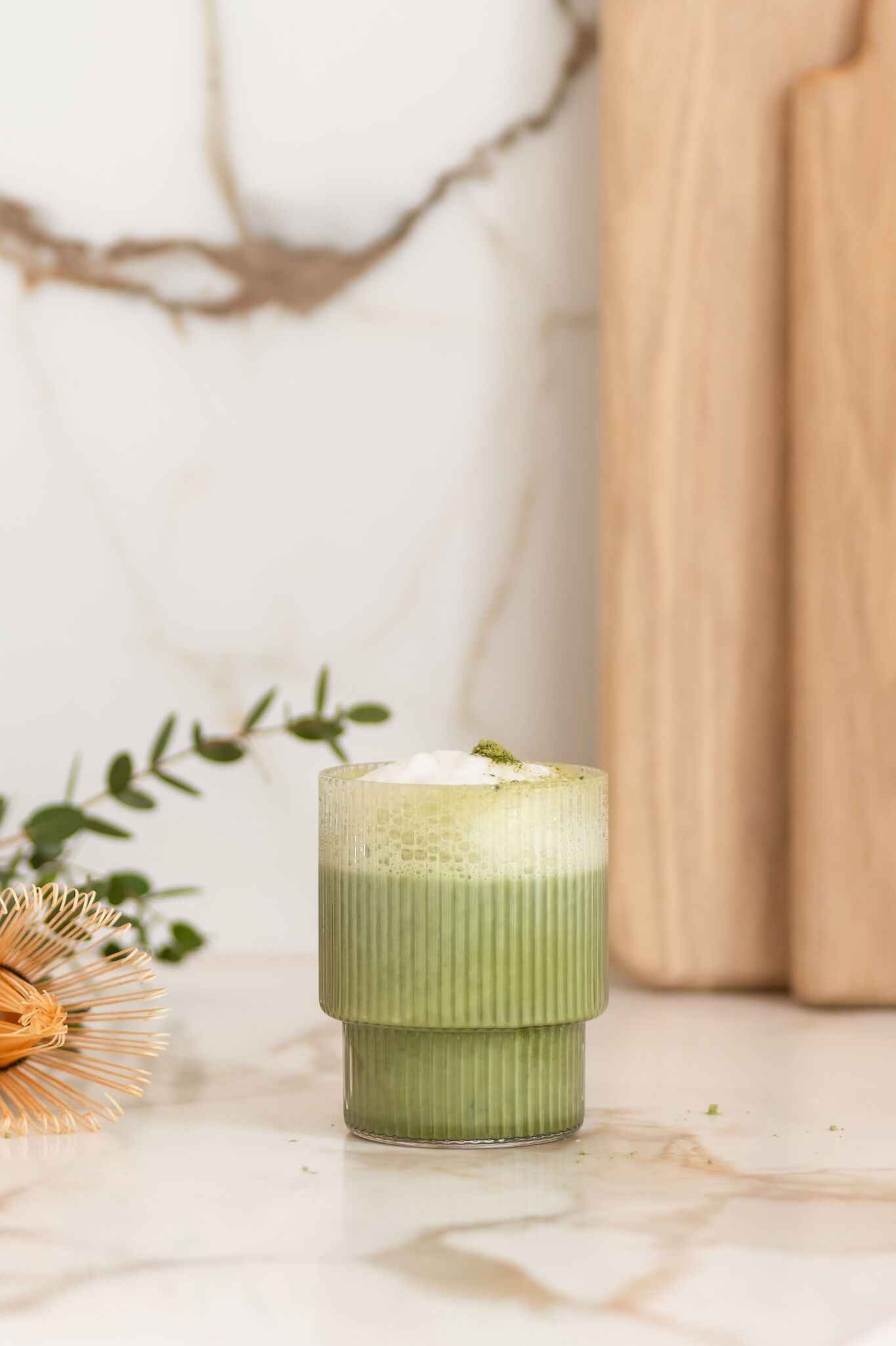 Green iced matcha latte with milk foam in a ribbed glass, styled on a marble table