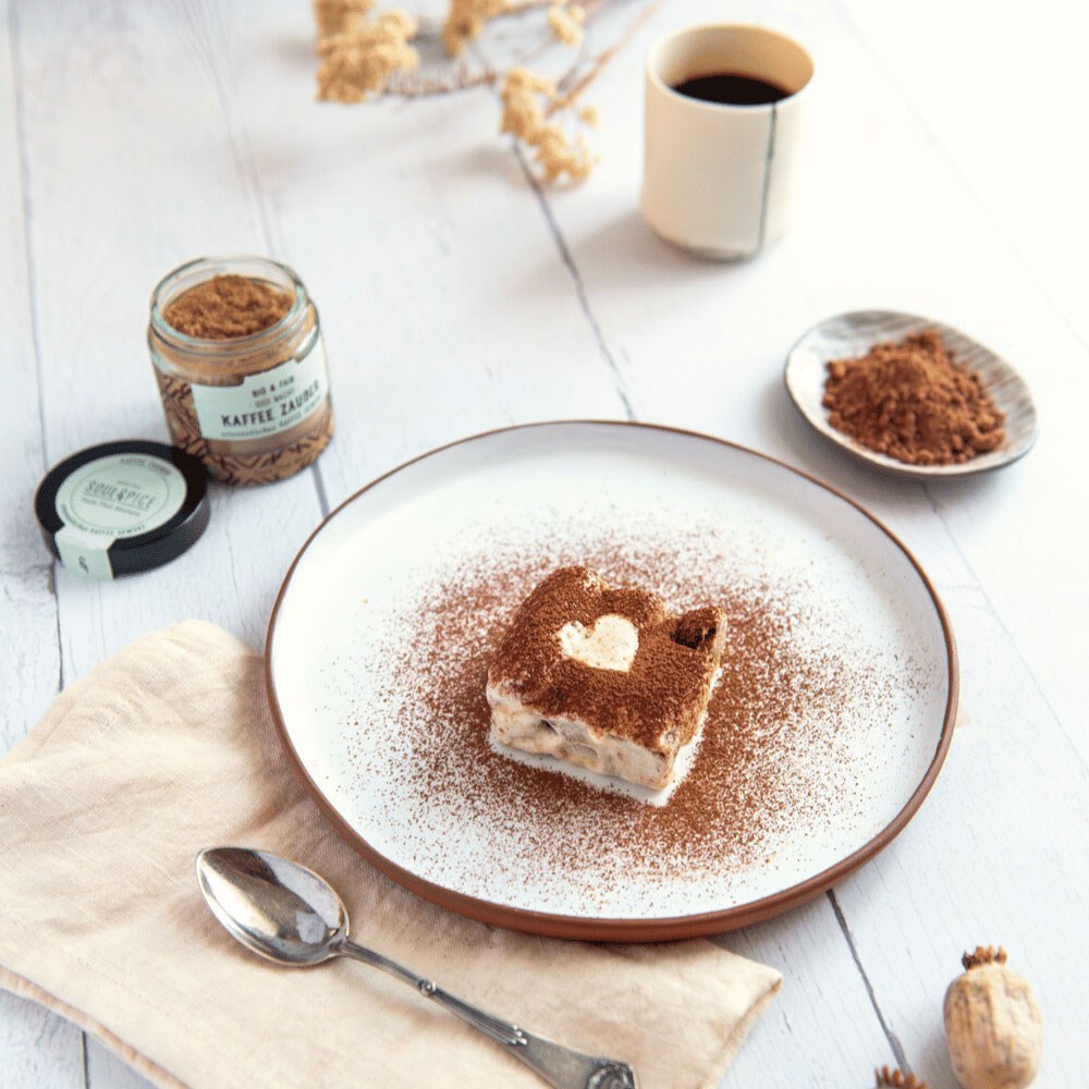 Quadratisches Dessert mit Kakao und Herzdekor auf weißem Teller, daneben Löffel, Tasse Kaffee und Glas Kaffeegewürz auf Holztisch, tiramisu, nachtisch, kaffee, puderzucker, teller