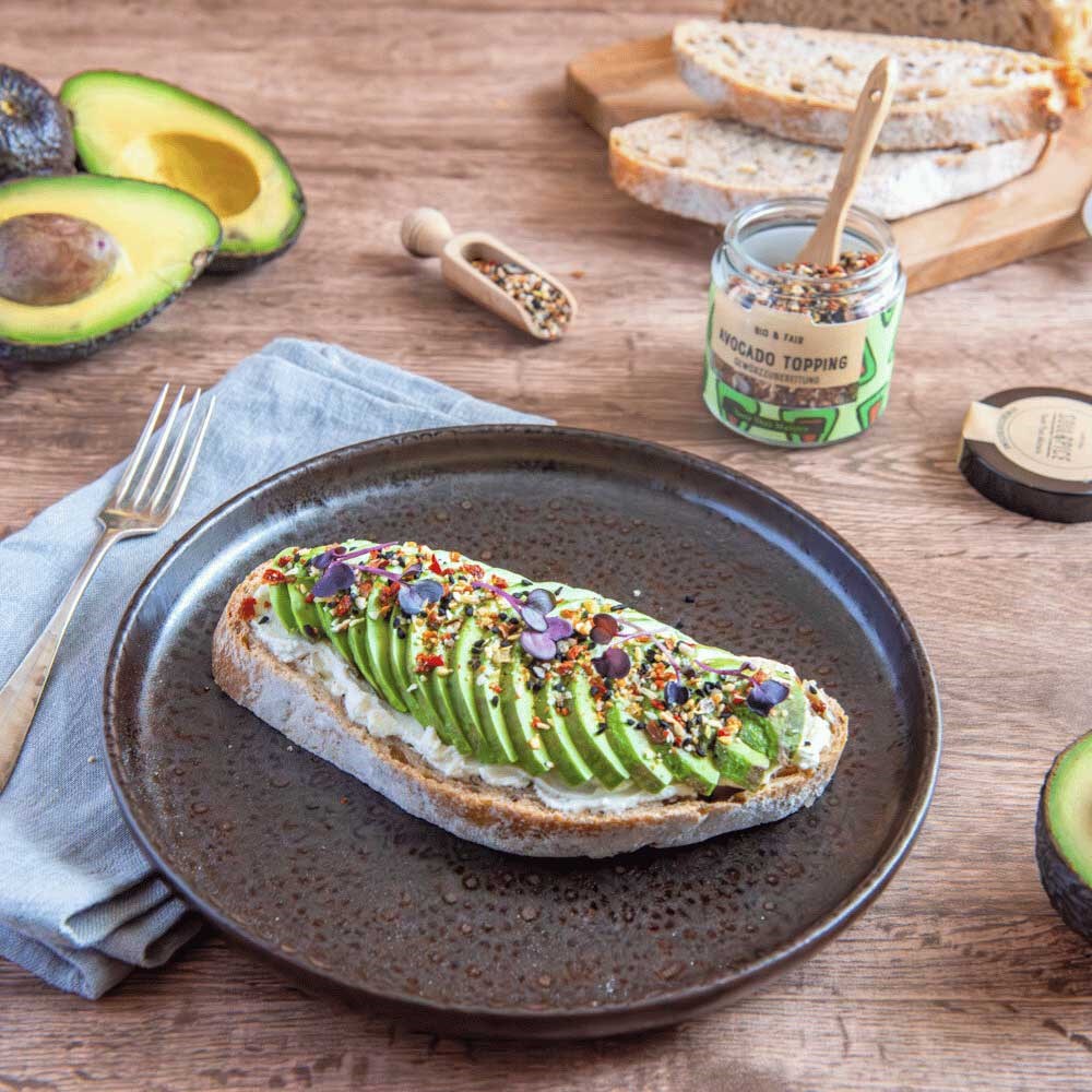 Slice of rustic sourdough topped with avocado slices, organic avocado seasoning and cress on ceramic plate, with fresh avocados and spice jar beside it