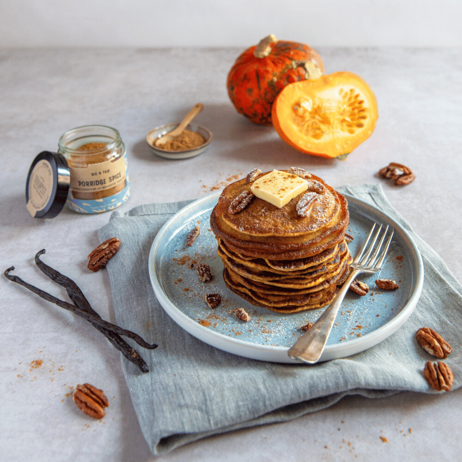 Stack of pumpkin pancakes with butter, pecans and spices on a plate, with vanilla pods, spice jar and fresh pumpkin in the background