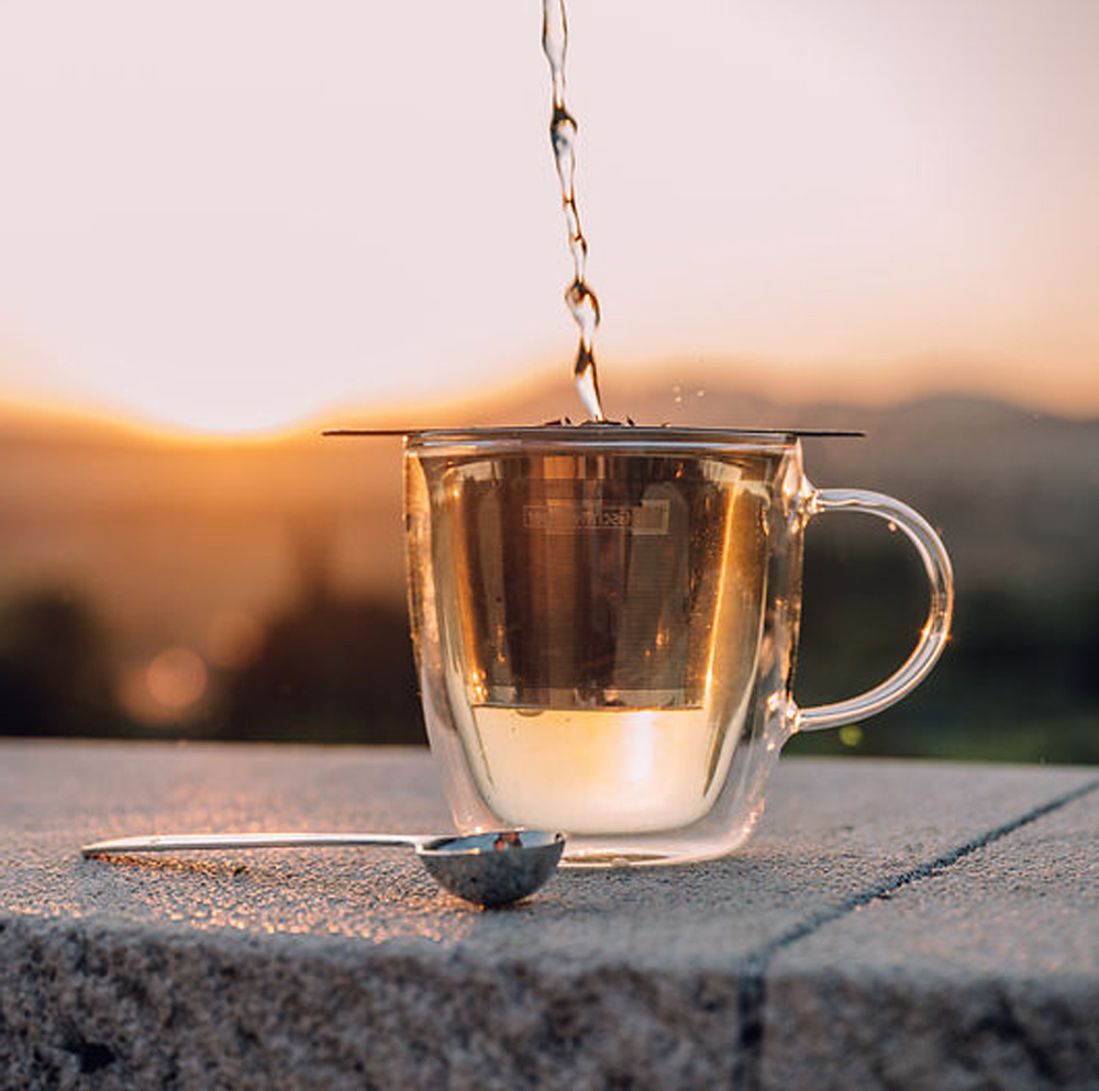 Heißes Wasser wird in eine doppelwandige Glasteetasse mit Edelstahlsieb gegossen, daneben liegt ein Teelöffel auf Stein, Sonnenuntergang im Hintergrund