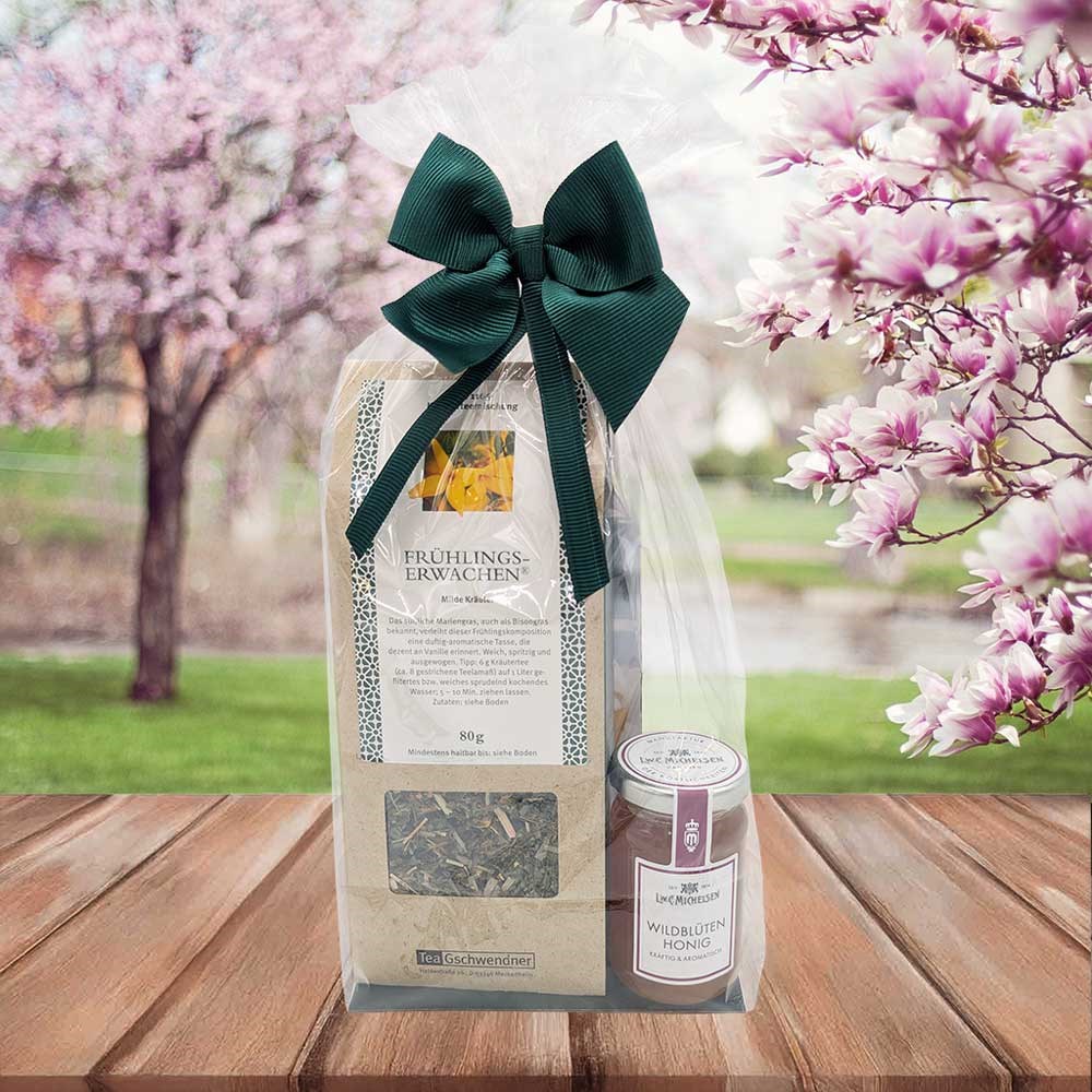 Geschenkset Frühlings­erwachen Kräutertee und Wildblütenhonig im Klarsichtbeutel mit grüner Schleife vor blühenden Frühlingsbäumen