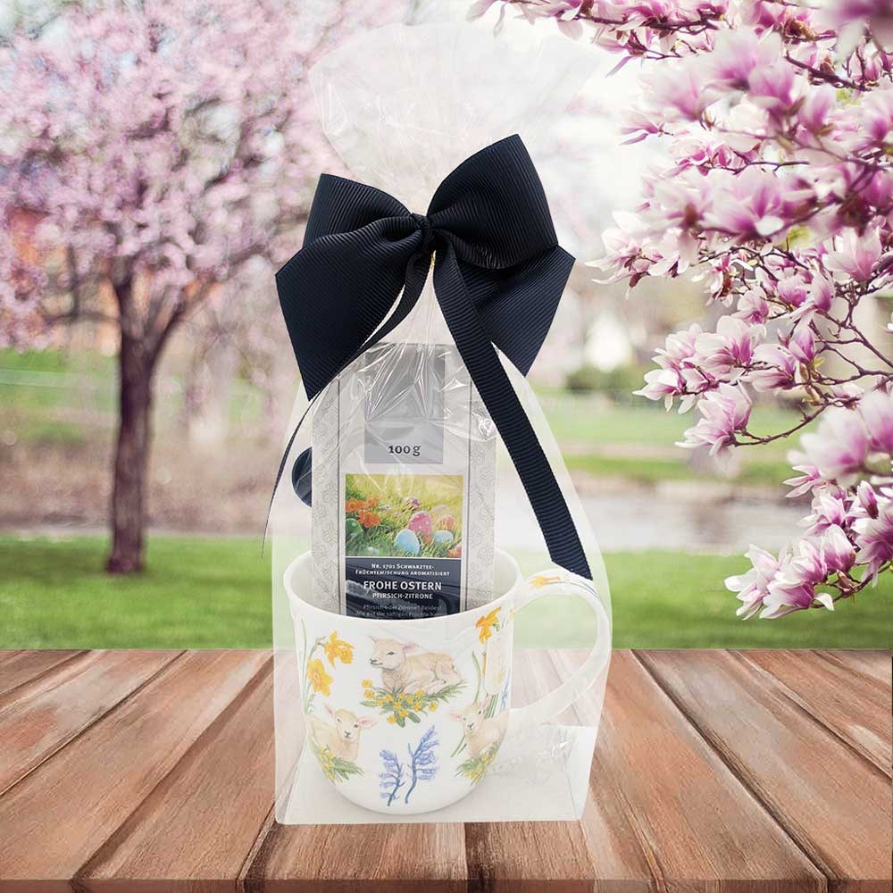 Oster-Geschenkset mit Frühlingsporzellantasse und Teepackung in Folie mit großer schwarzer Schleife vor blühenden Bäumen