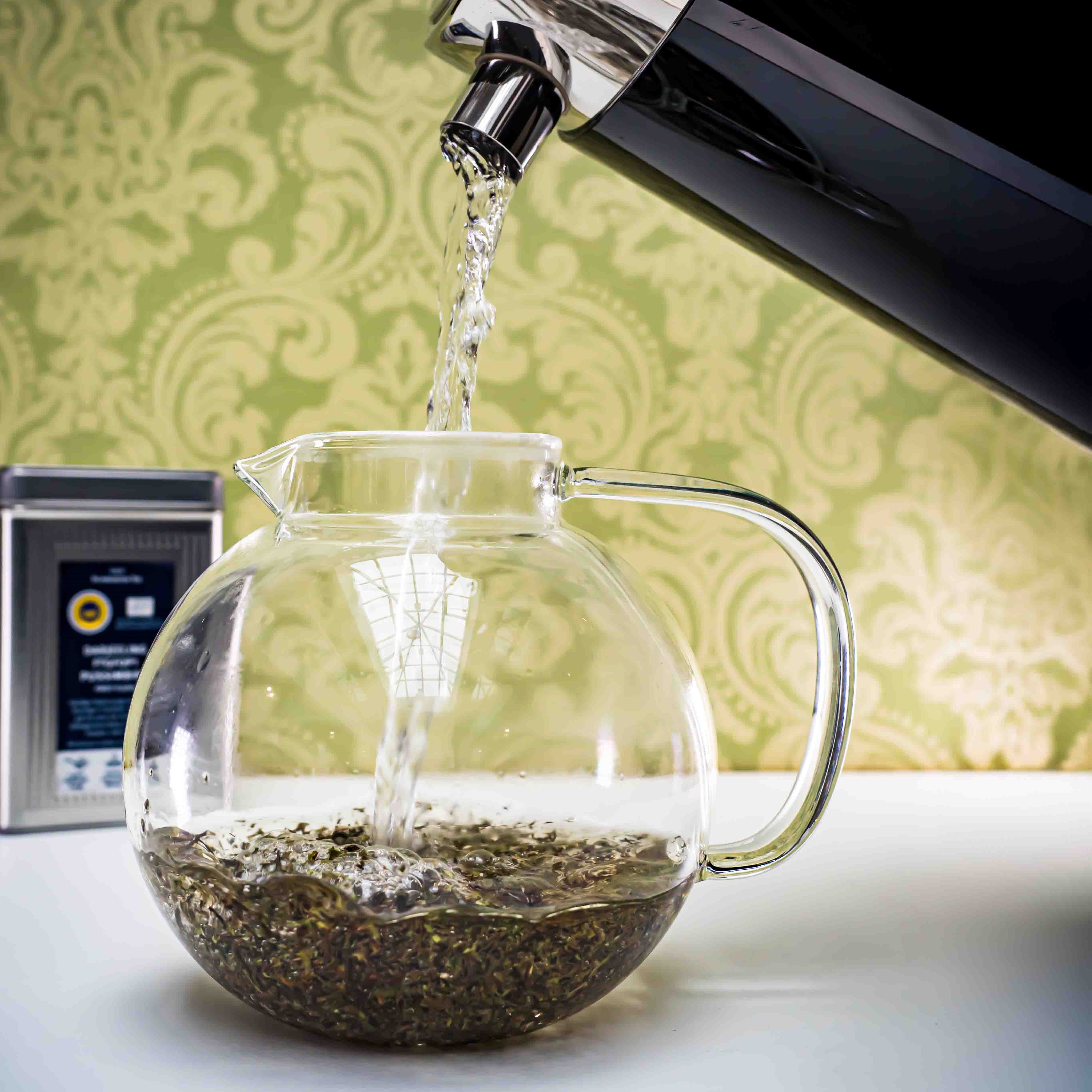 Heißes Wasser fließt in eine runde Glasteekanne mit Tee, Glaskanne, Kaffeekocher, gläserne Karaffe, Kaffee-Kanne, Filterkaffee